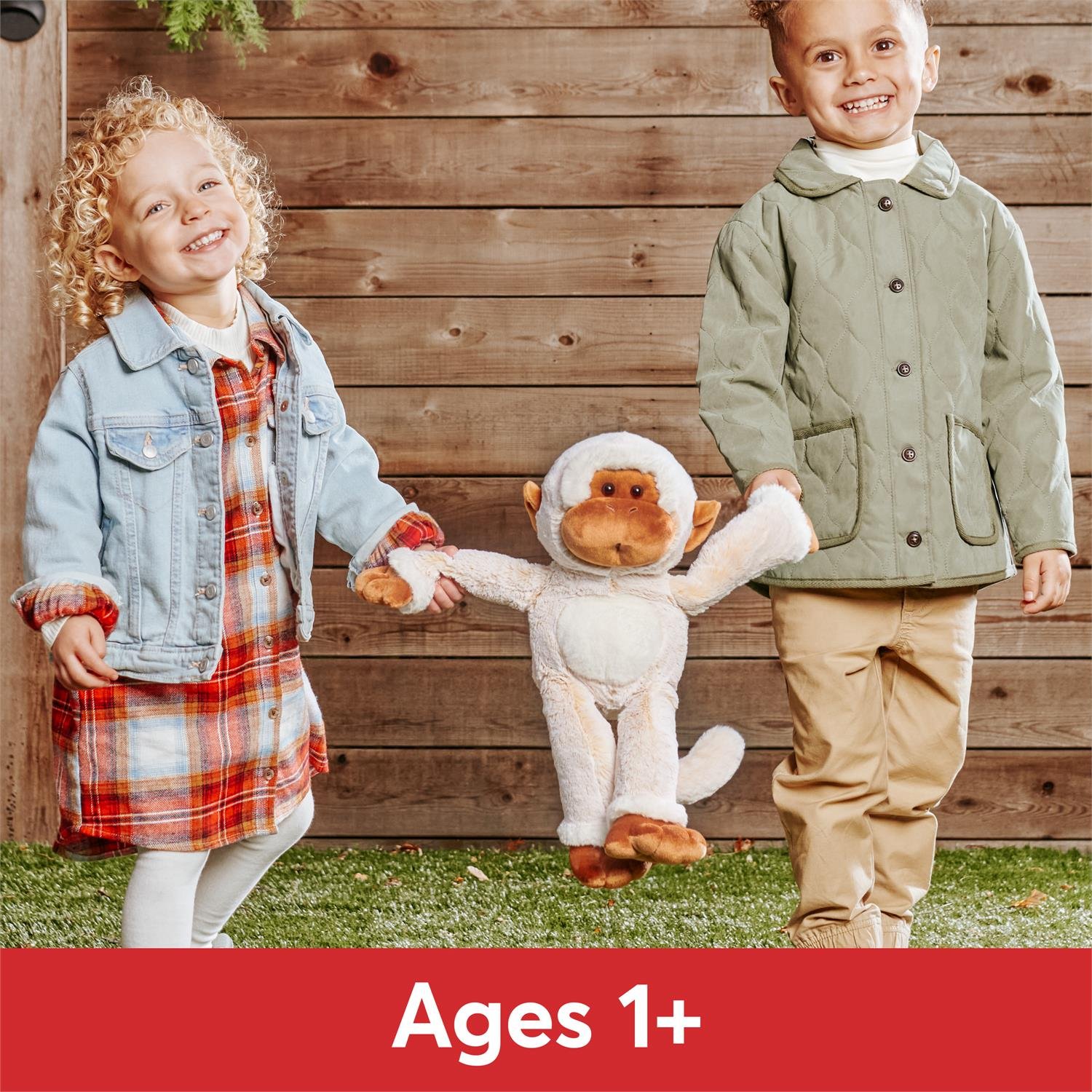 Two children holding a stuffed monkey toy with a wooden fence background and 'Ages 1+' label at the bottom.