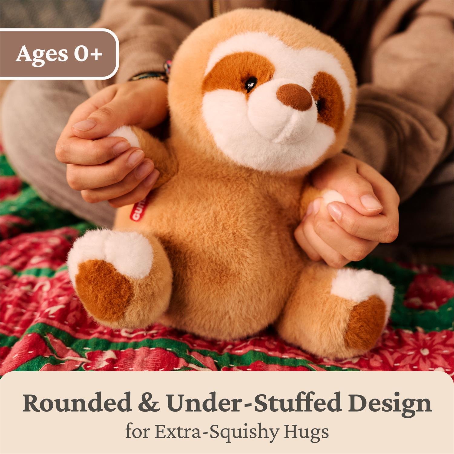 A plush sloth toy being held in both hands over a red patterned blanket, with a person's torso and legs visible in the background. Text overlays read 'Ages 0+' and 'Rounded & Under-Stuffed Design for Extra-Squishy Hugs'.