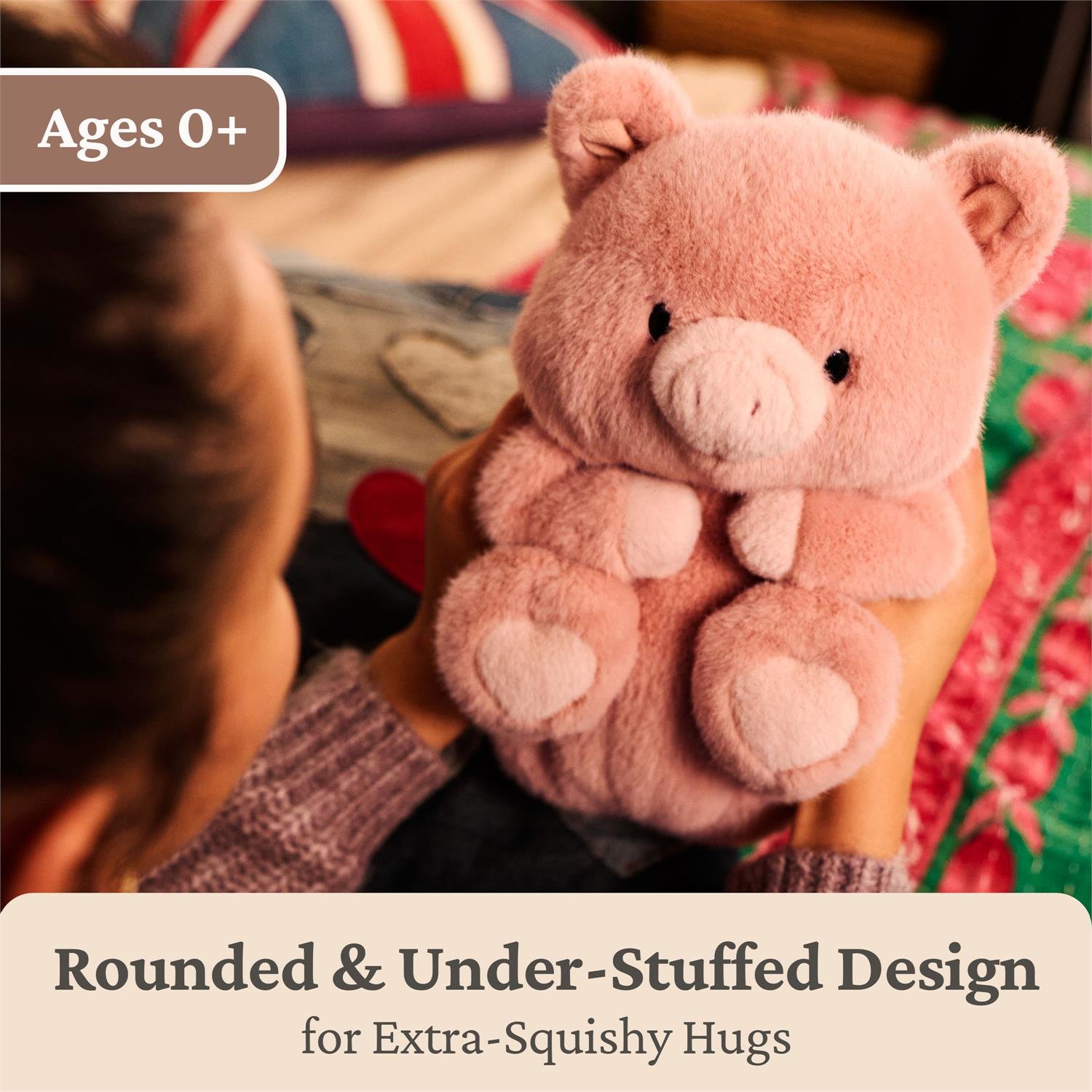 A pink plush teddy bear being held by a person in a cozy indoor setting, with overlaid text reading 'Ages 0+' and 'Rounded & Under-Stuffed Design for Extra-Squishy Hugs.' The person is partially visible from behind, and the bear is the clear product focus.