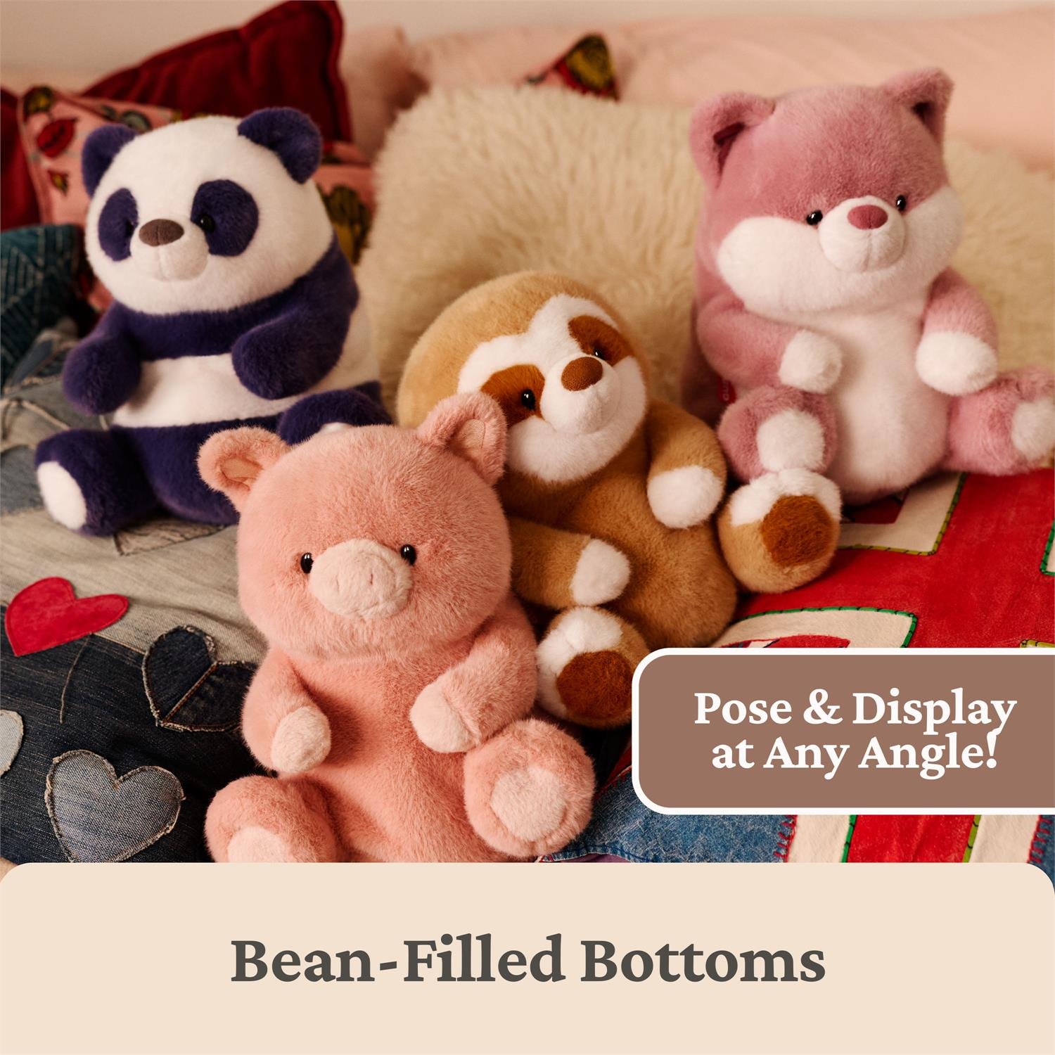 Four plush stuffed animals arranged together on a quilted blanket with pillows: a panda, a sloth, a pink fox, and a pink bear. A brown label says 'Pose & Display at Any Angle!' and text below reads 'Bean-Filled Bottoms.