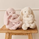 Two plush bunnies, one pink and one white, hugging smaller versions of themselves, sitting on a weathered wooden stool.