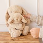 Two plush tan bunnies held by a child wearing a beige speckled sweater, sitting on a wooden surface.